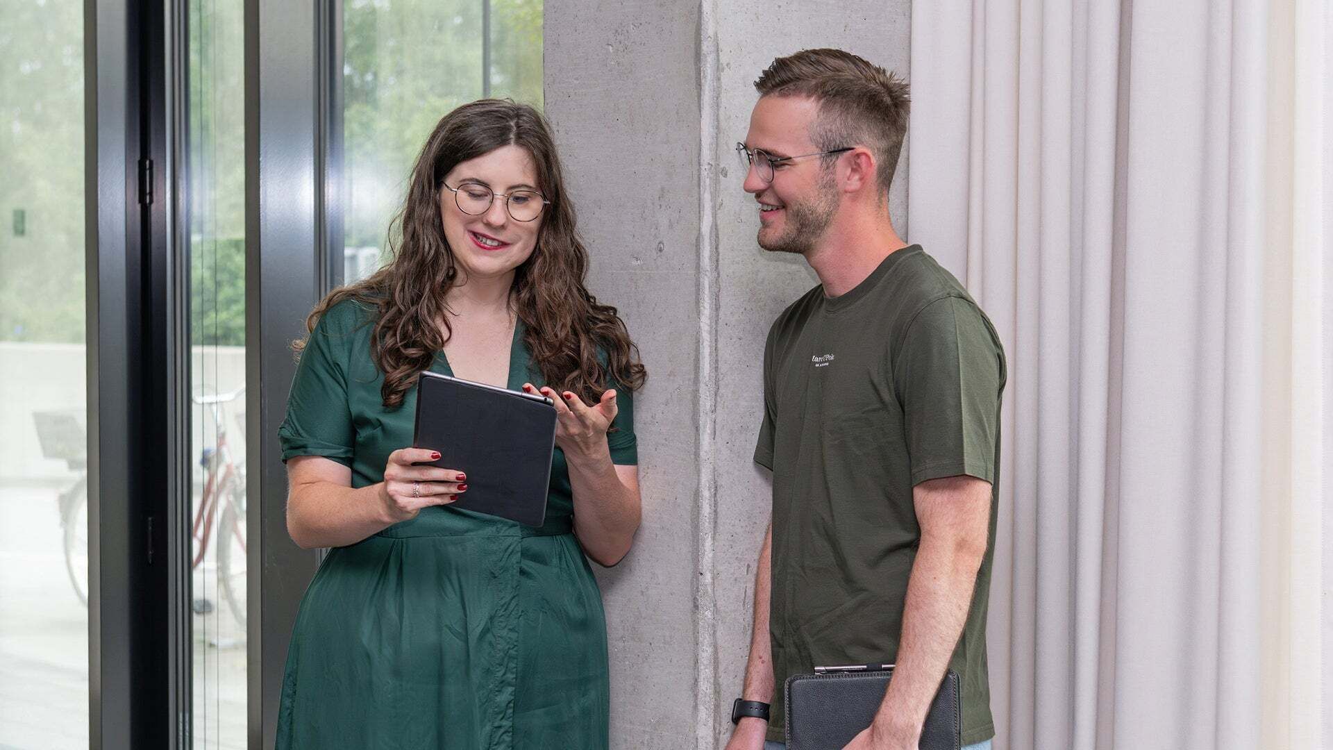 Ein Mann und eine Frau stehen vor einer Betonsäule und schauen auf ein Tablet, welches die Frau in der Hand hält