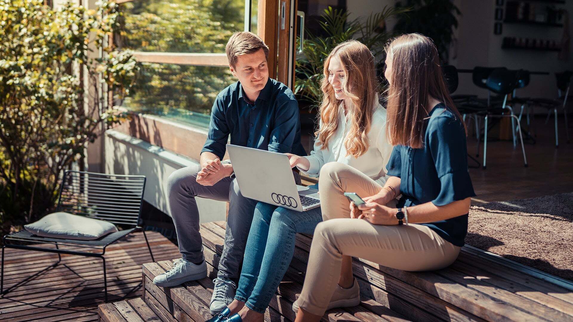 Drei Personen sitzen draußen bei Sonnenschein mit Laptop auf dem Schoß und reden.