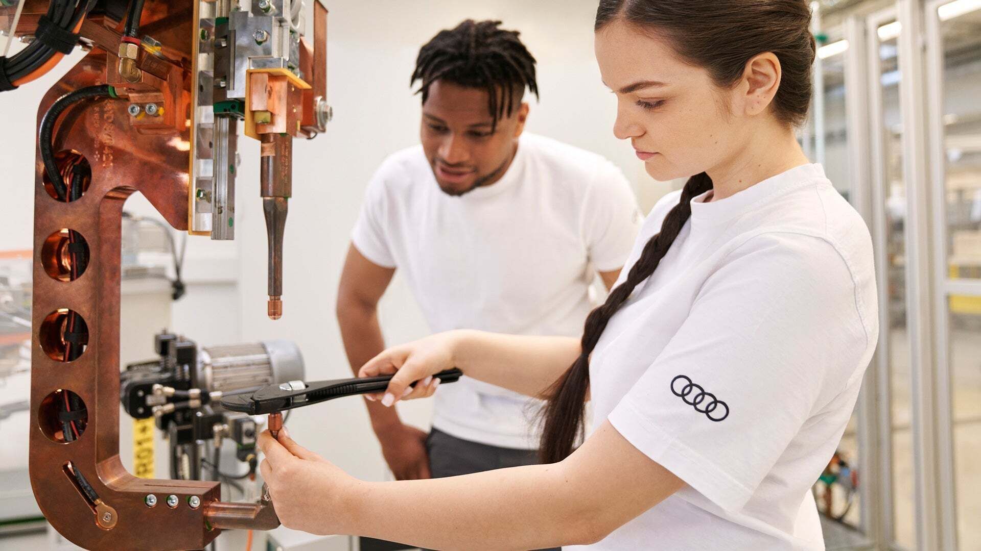 Zwei Audi Mitarbeitende stehen an einer industriellen Maschine. Während die eine Person mit einem Werkzeug an der Vorrichtung arbeitet, beobachtet die zweite Person den Arbeitsvorgang.