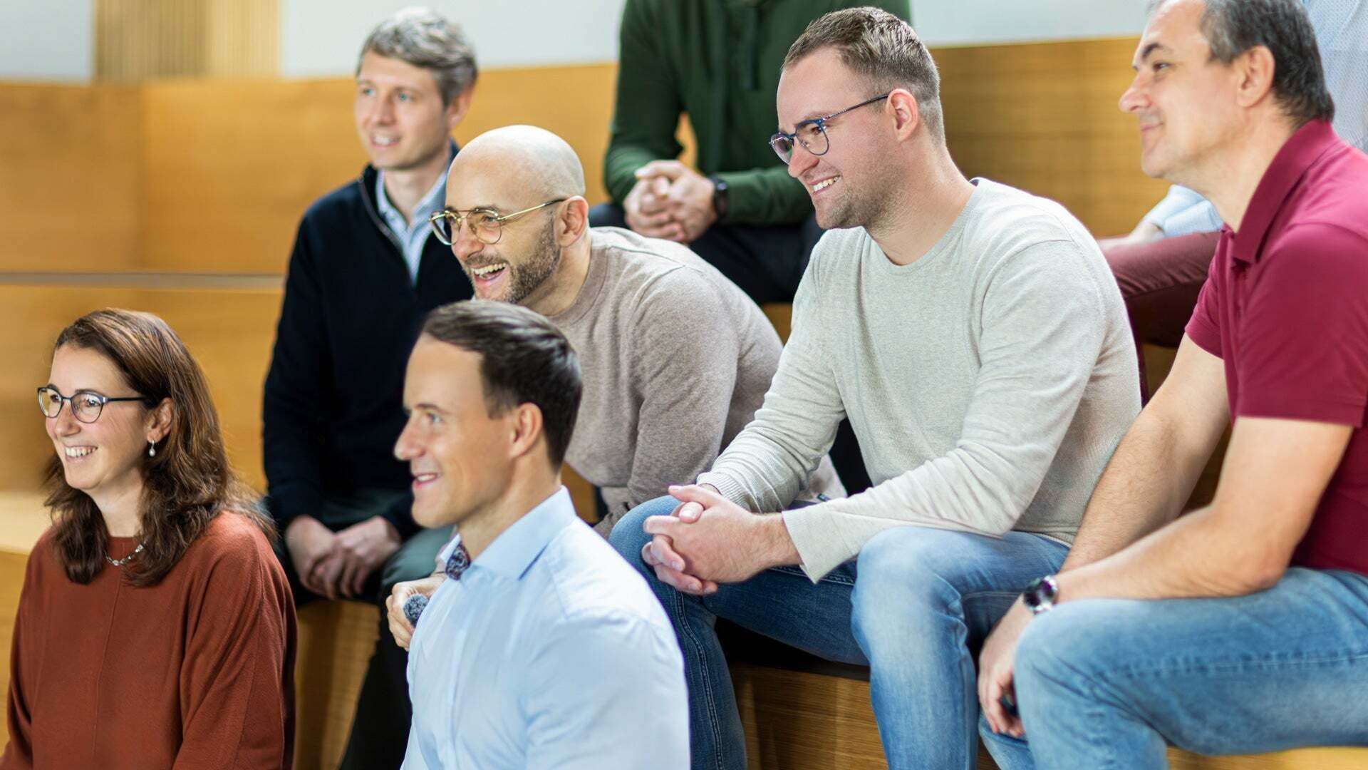 Gruppe von Audi Mitarbeitenden sitzt auf Holzstufen in einem hellen Raum und blickt gemeinsam nach vorn.