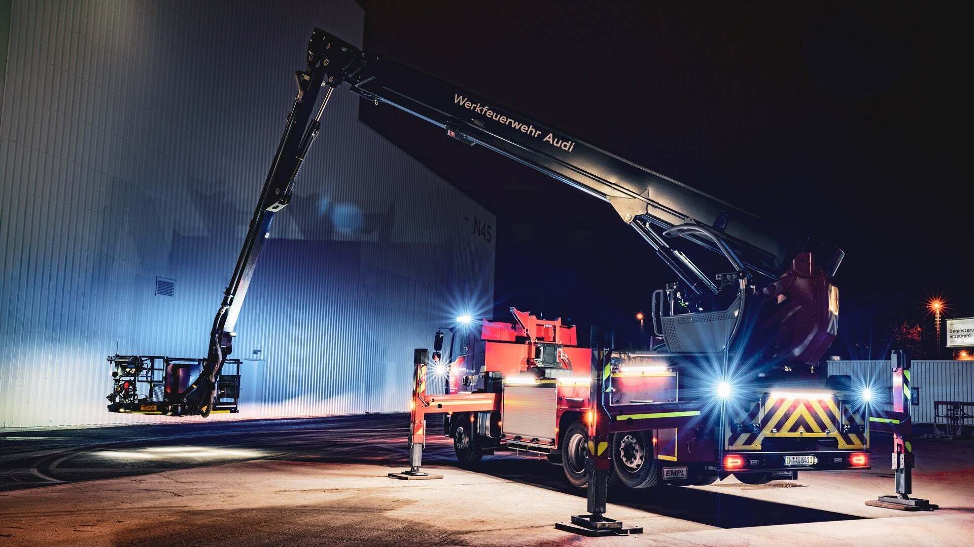 Ein Feuerwehrfahrzeug mit ausgefahrenem Teleskopmast trägt die Aufschrift Werkfeuerwehr Audi und beleuchtet eine große Hallenwand bei Nacht.