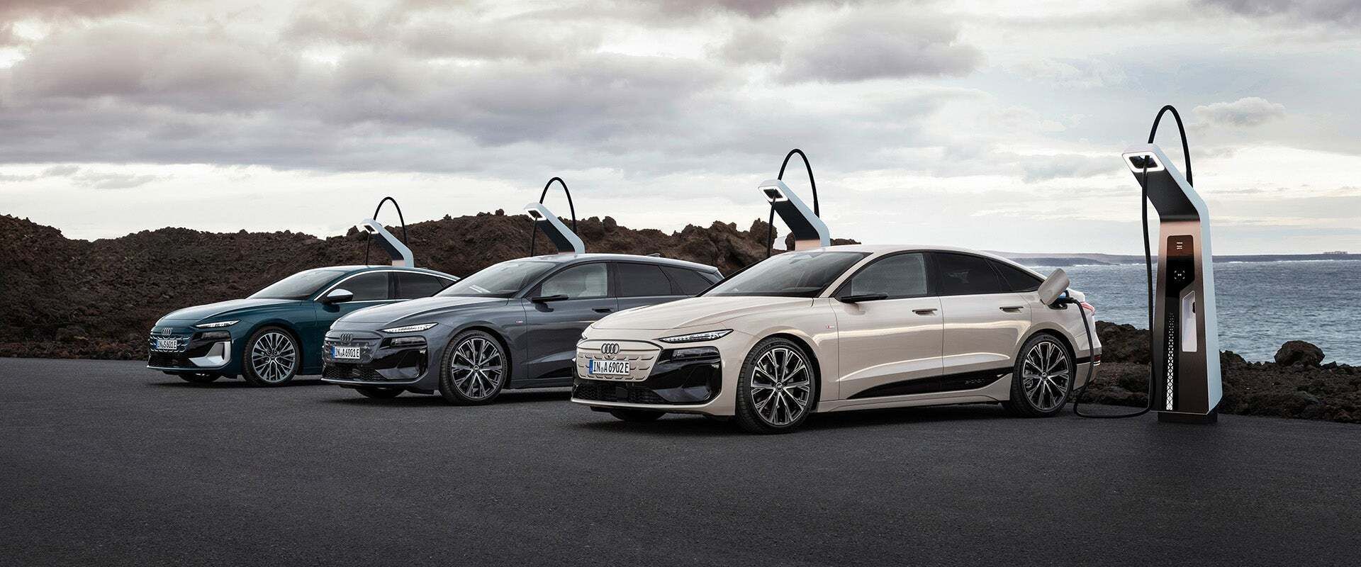 Drei Audi A6 e-tron Fahrzeuge in verschiedenen Farben stehen an einer Ladestation vor einer felsigen Küstenlandschaft mit bewölktem Himmel. Das vorderste Fahrzeug ist an die Ladesäule angeschlossen.