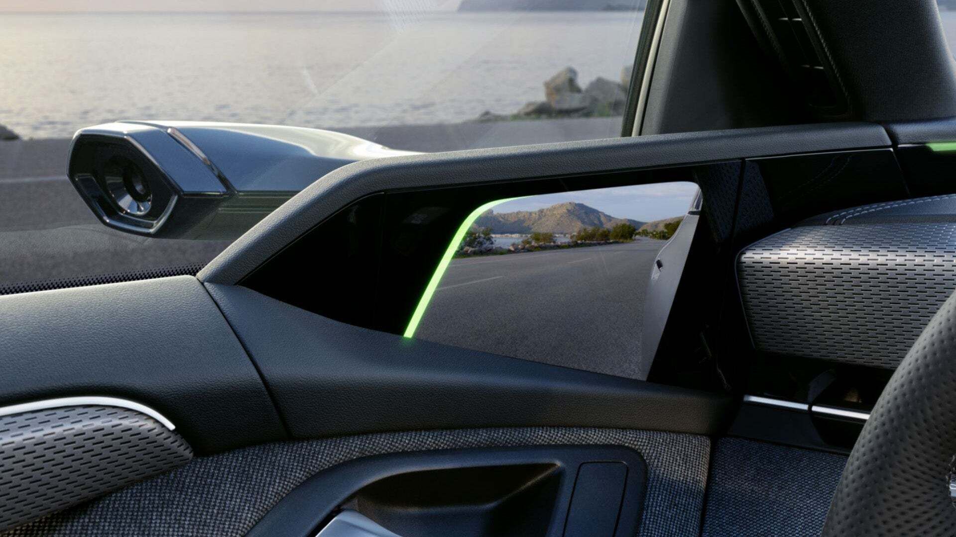 Detailansicht eines digitalen Außenspiegels in einem Audi. Die Kamera auf der Fahrzeugaußenseite überträgt das Bild auf einen Bildschirm im Innenraum. Im Hintergrund ist eine Küstenstraße mit Blick auf das Meer und die Berge zu sehen.