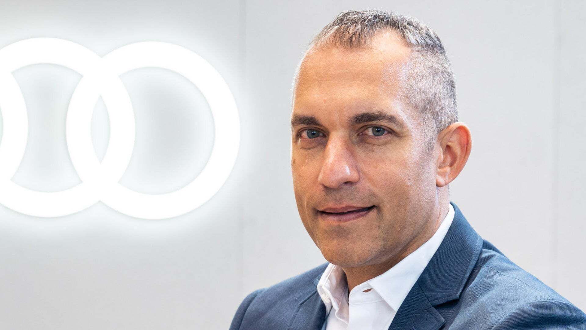 Mann im Anzug (Daniel Patnaik) vor beleuchtetem Audi-Logo, Blick in die Kamera.