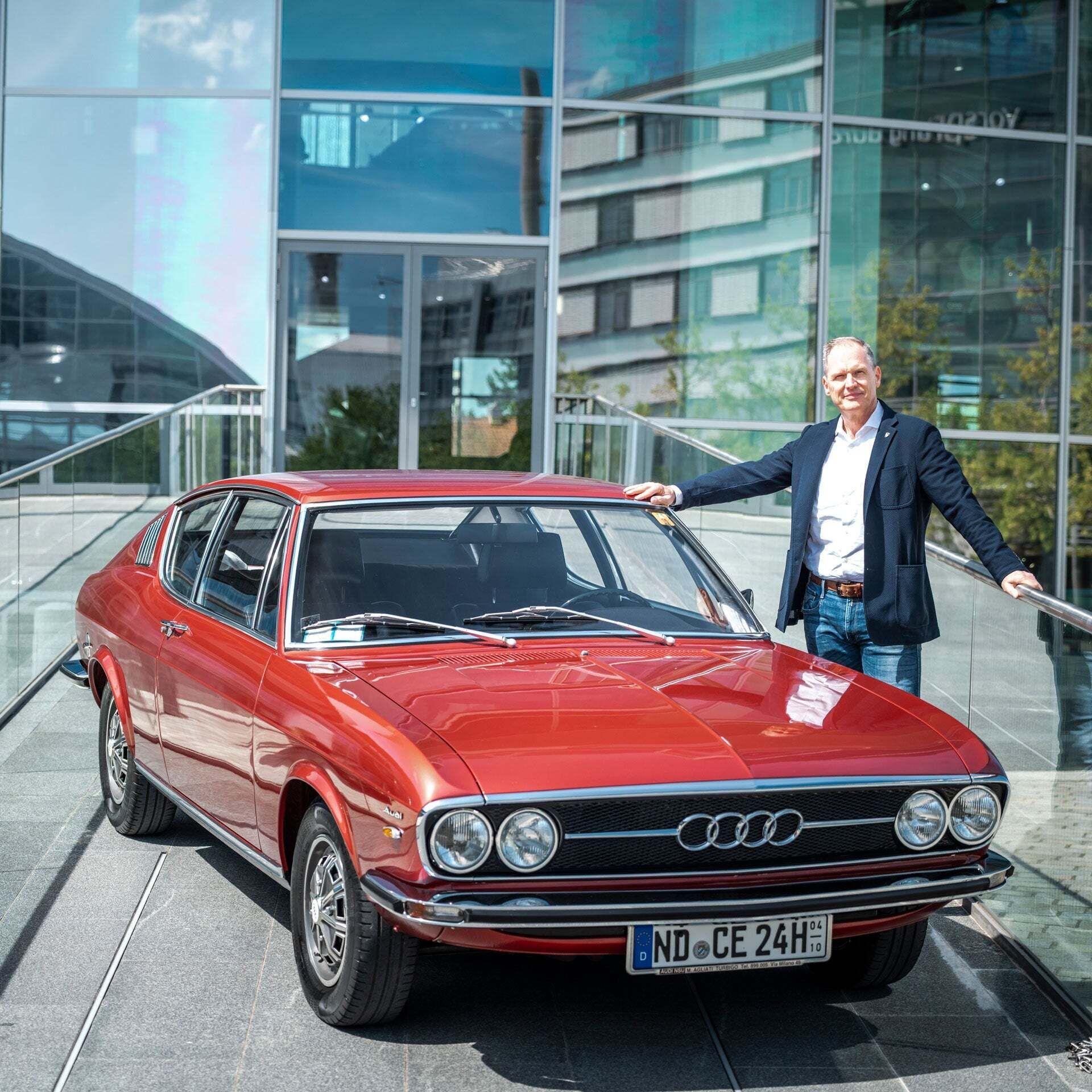 Rene Marc Heinze in a dark jacket, white shirt and jeans leans against his red Audi 100 on the ramp of the Audi museum mobile