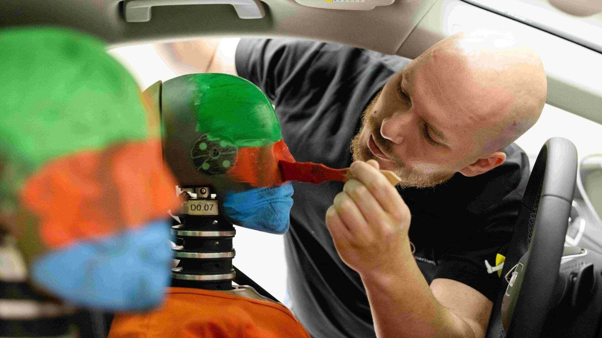 Ein Mann pinselt rote Farbe an den Kopf eines Crashtest-Dummies, der auf dem Fahrersitz eines Testfahrzeugs sitzt. 