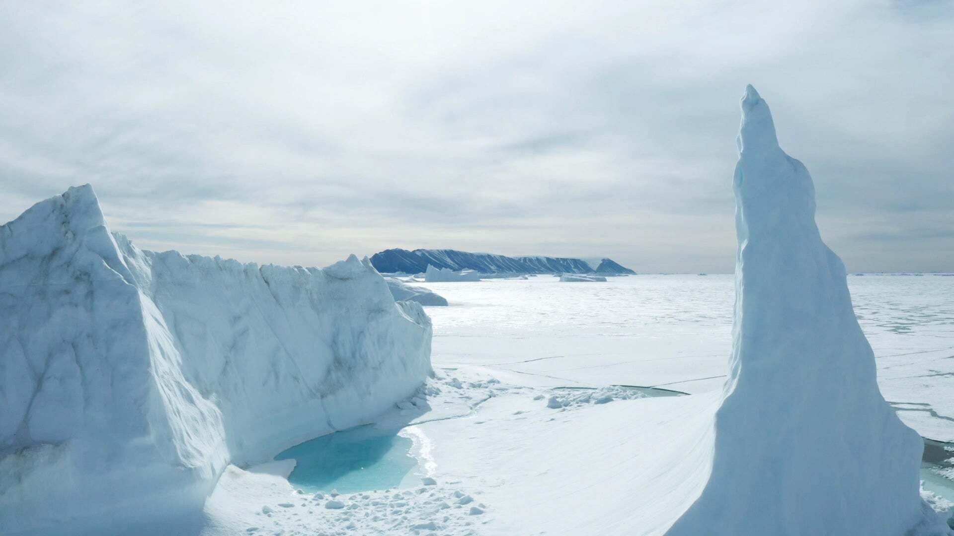 Icy landscape with rugged glacier, frozen sea and mountains in the background under a cloudy sky.