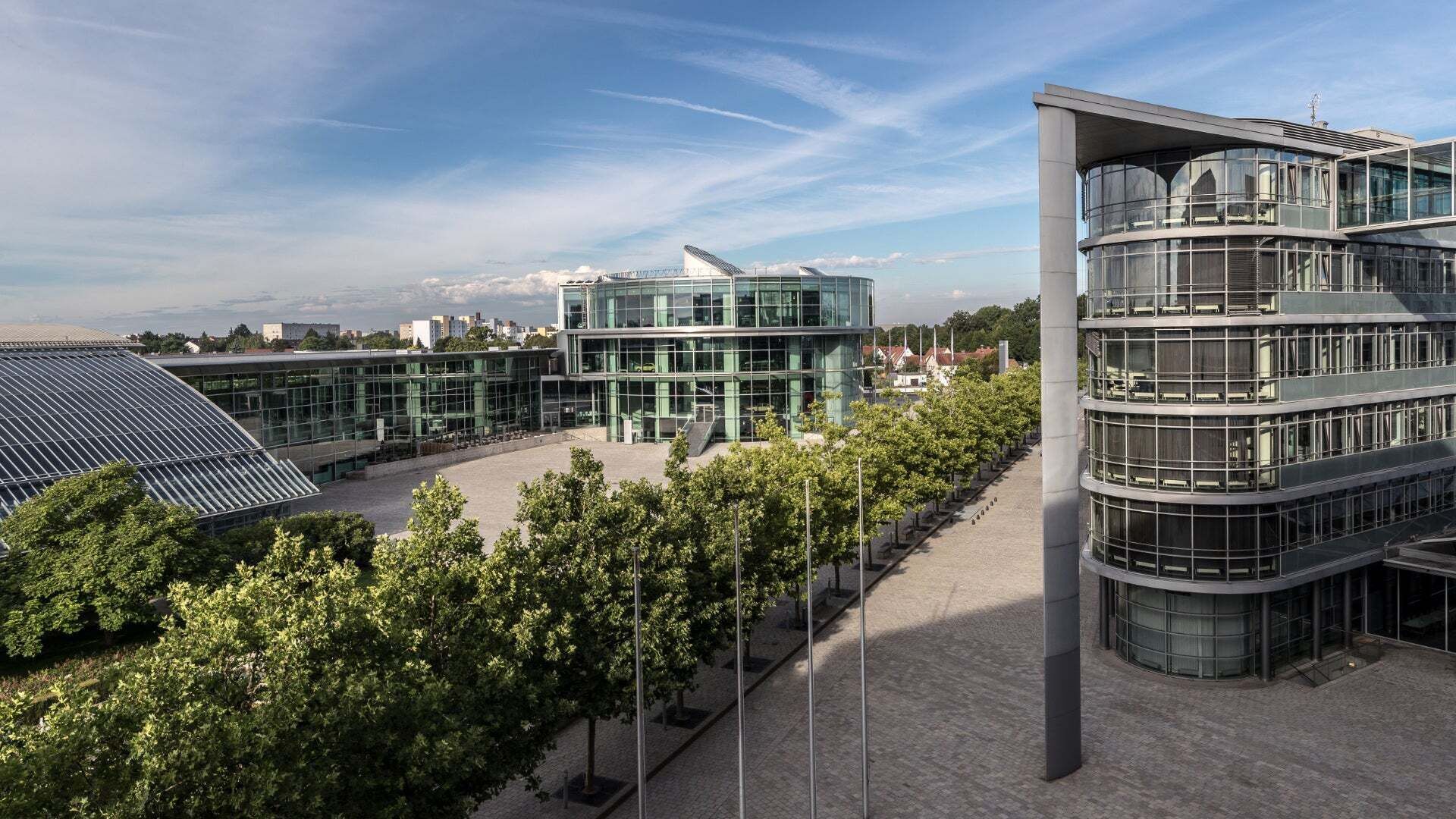 Ein moderner Gebäudekomplex mit großen Glasfassaden und geschwungenen Formen, umgeben von einer Baumreihe und gepflasterten Wegen unter einem klaren blauen Himmel. 