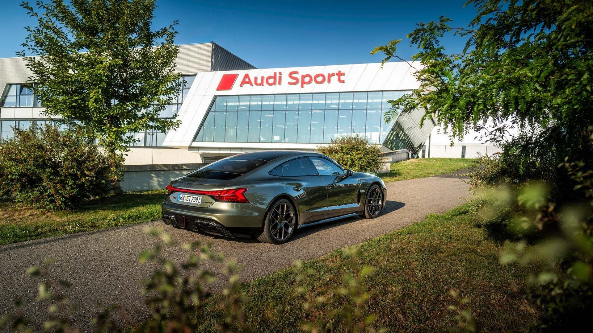 A green Audi RS e-tron GT on a driveway, with the company headquarters bearing the Audi Sport GmbH logo in the background.