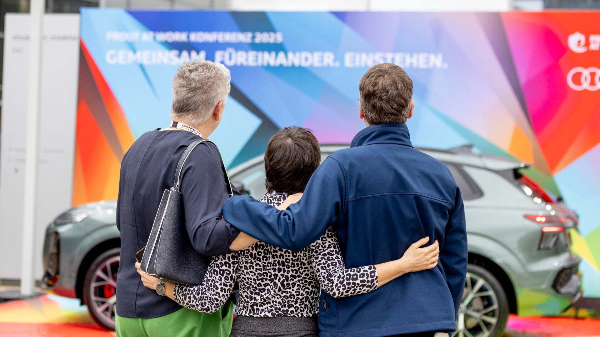 Standing together for diversity and equal opportunities – employees stand arm in arm, looking at a poster on the topic of Audi Diversity.