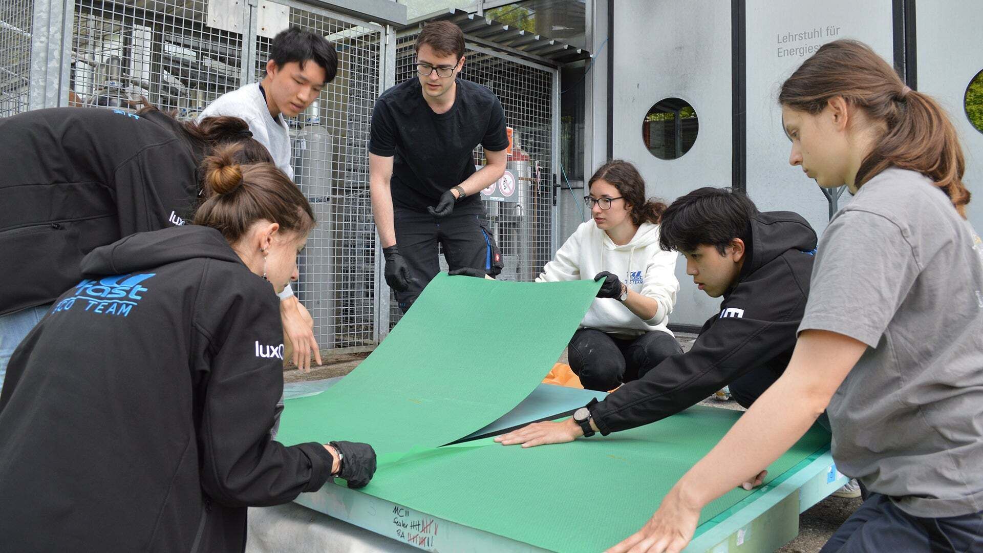 A few people sit and stand around a green cardboard on the floor