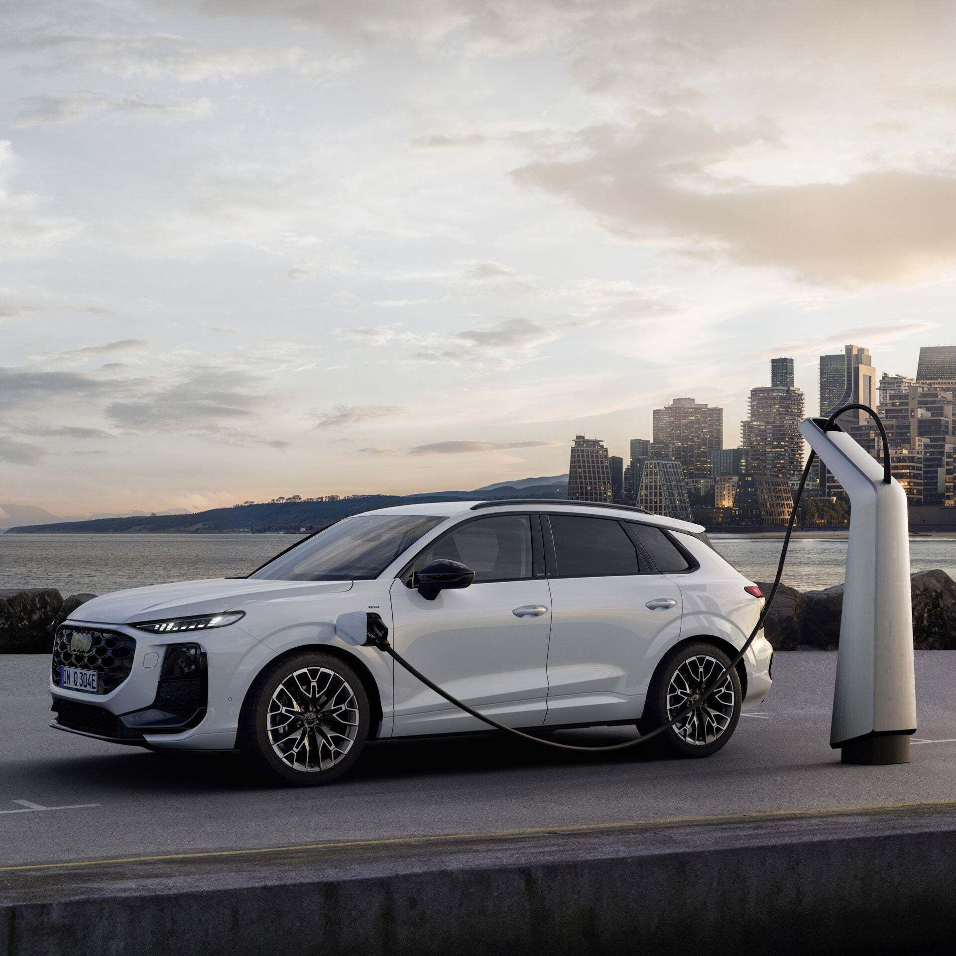 White Audi Q3 SUV e-hybrid at a silver charging station in front of a city backdrop with skyscrapers.