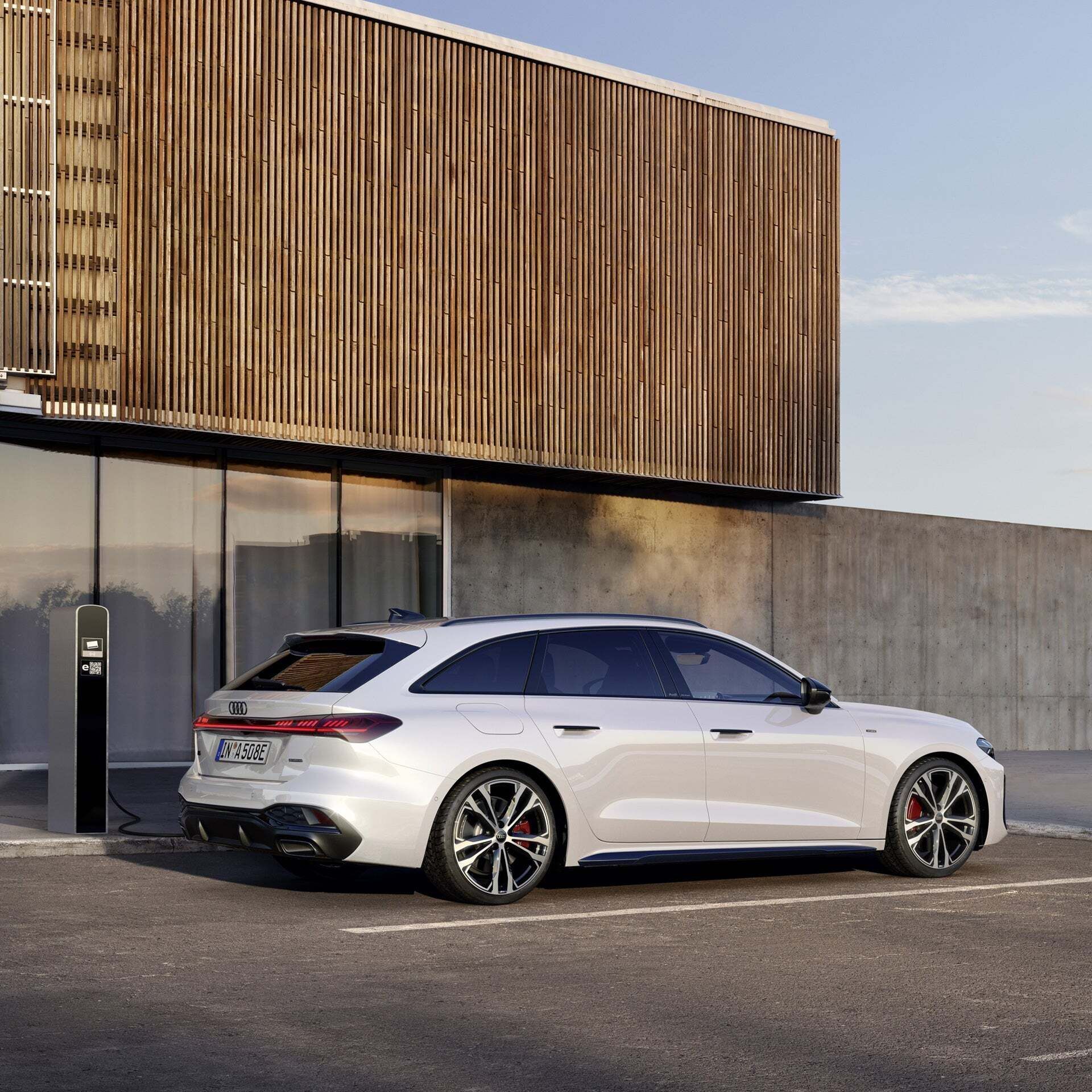 A white Audi A5 Avant e-hybrid charges at a charging station in front of a modern building with a façade of glass, concrete and wooden panels.