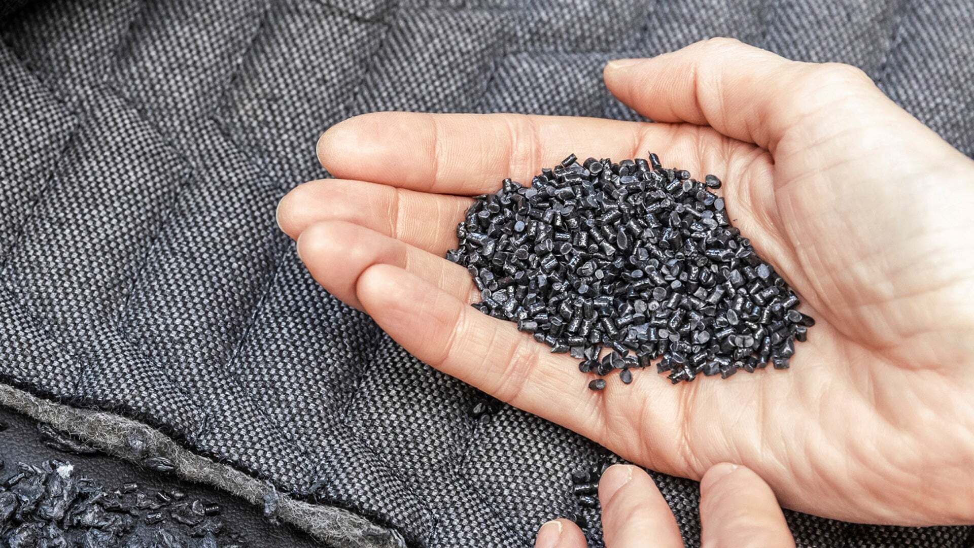 Hand hält dunkle Granulatkörner über einem dunklen textilen Material, umgeben von weiterem Granulat.