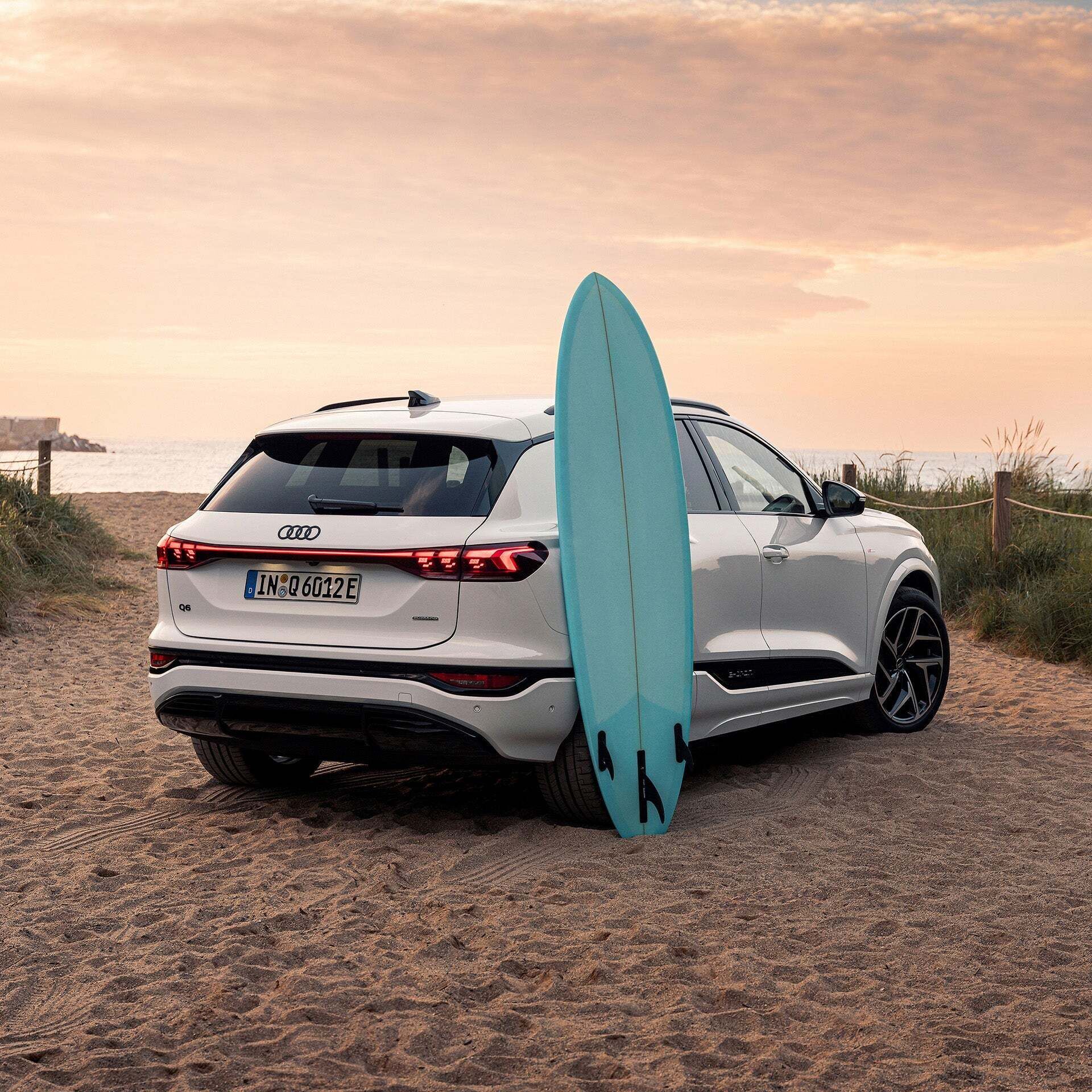 A white Audi Q6 SUV e-tron is parked at the beach crossing, with the sea in the background. A turquoise surfboard is leaning against the rear side door.