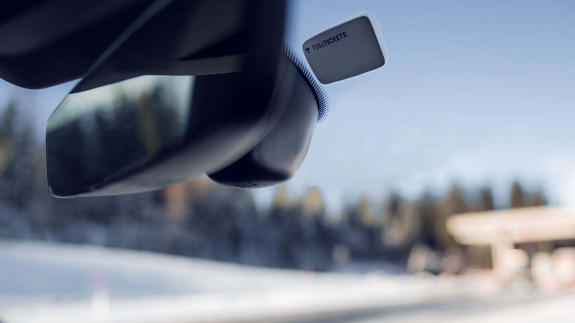 A small white toll box is stuck to the windscreen to the right behind the rear-view mirror. There is a toll station visible blurred in the background.