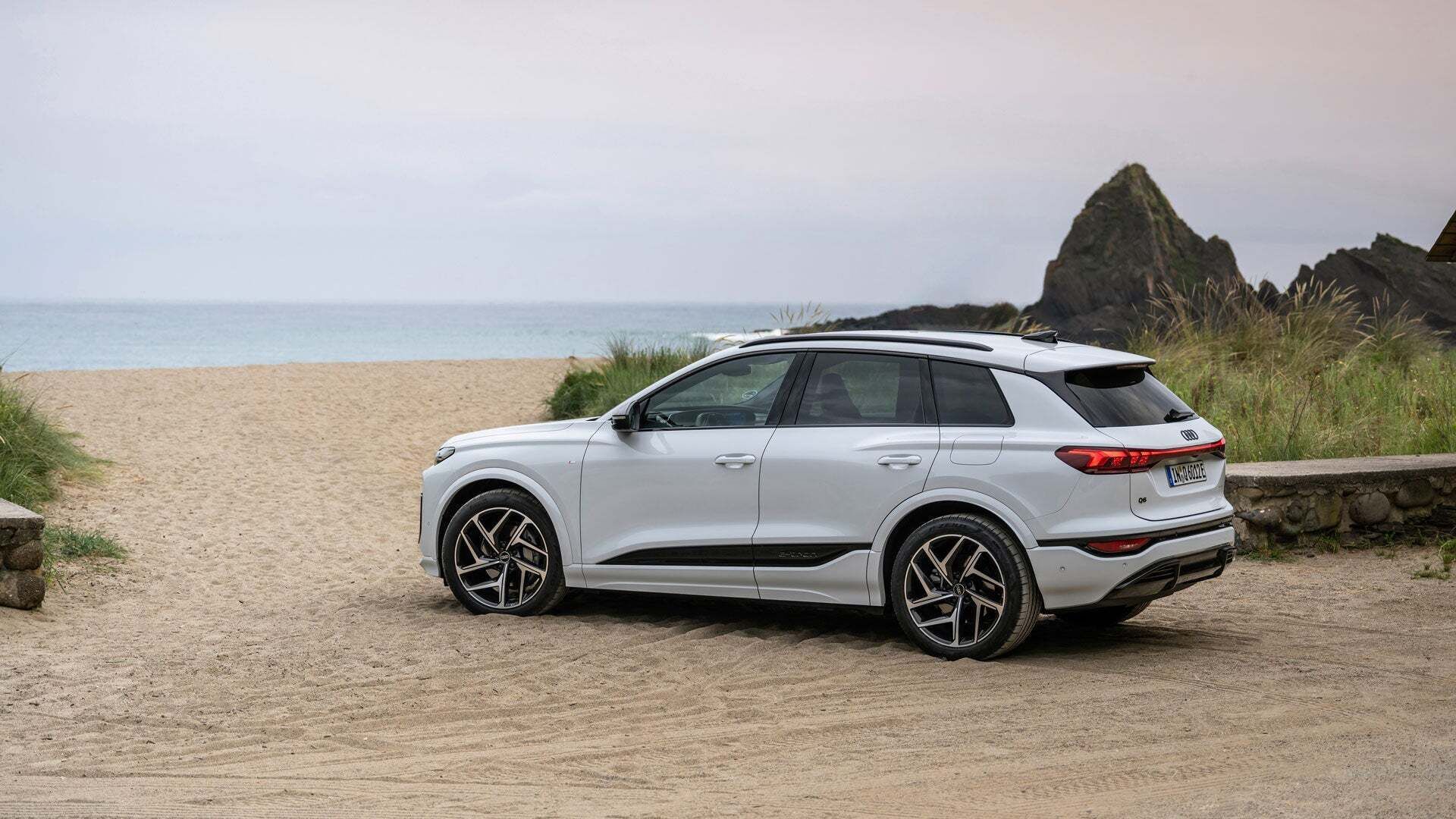 White Audi Q6 SUV e-tron at a beach crossing. Behind the vehicle is a wide sandy beach facing the open sea.