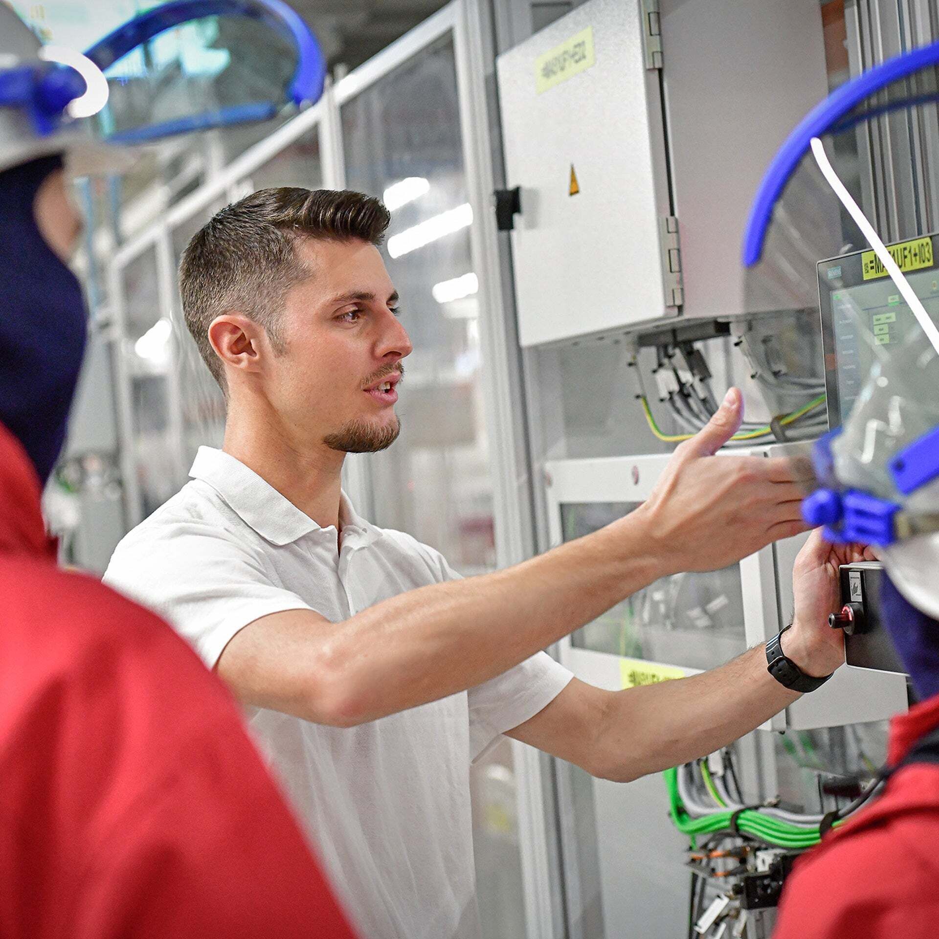 A man explains how to operate a machine control system in a production hall to two people wearing protective clothing.