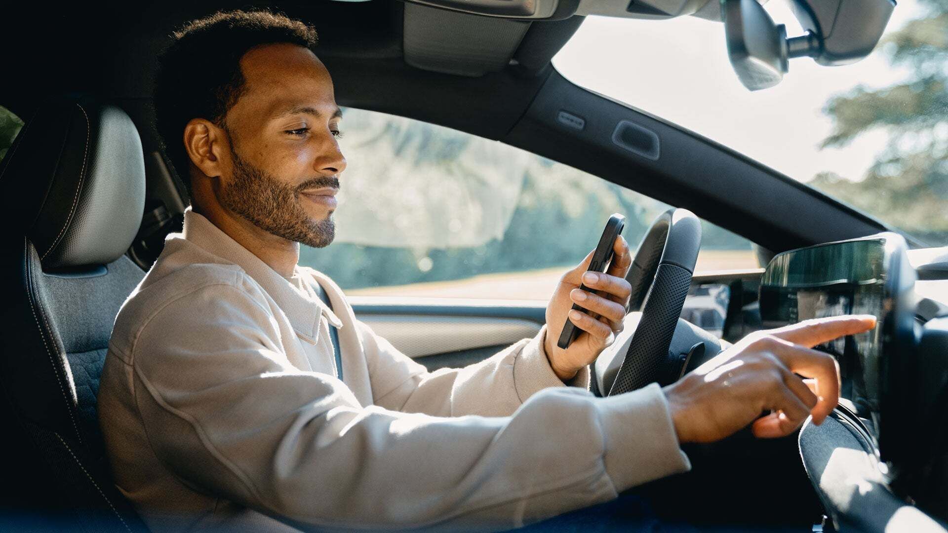 Mann sitzt hinter dem Steuer seines Audi. Er hält sein Smartphone in der einen Hand, während er mit der anderen Hand das MMI des Fahrzeugs bedient. 