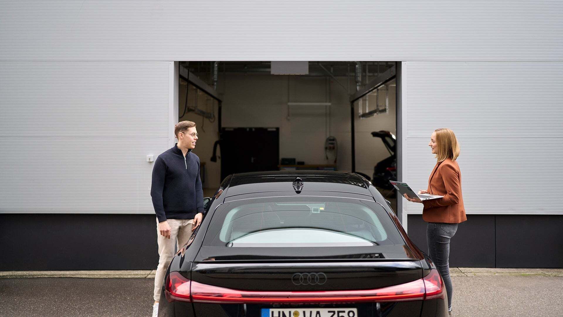 Sophia Schupritt steht ihrem Kollegen gegenüber und hat einen Laptop in der Hand. Es befindet sich ein Audi zwischen ihnen