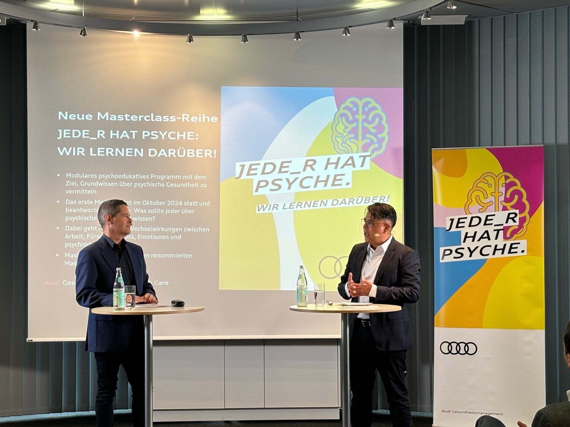 Zwei Männer sprechen auf Bühne bei Audi-Event zur Reihe 'Jede_r hat Psyche' mit Bannern und Präsentation im Hintergrund.
