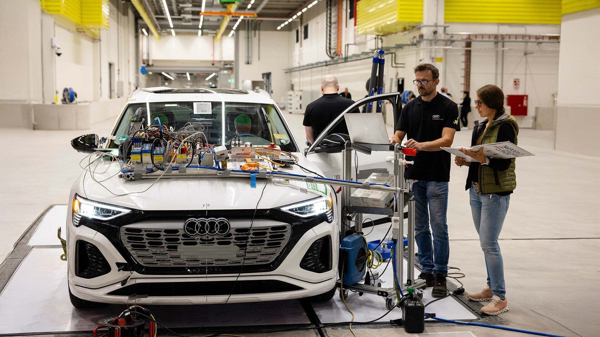 Weißer Audi mit Verkabelung und Testgeräten auf der Motorhaube in einer Werkstatt, während zwei Ingenieure die Daten prüfen.