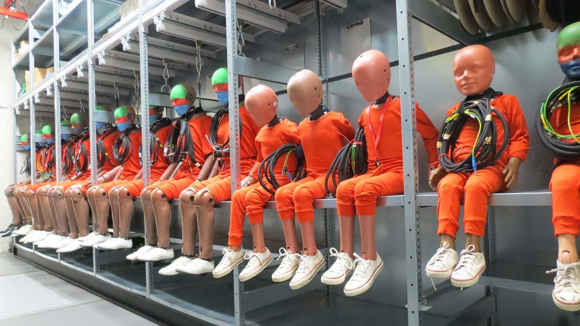 Eine Reihe von Crashtest-Dummys in orangefarbenen Anzügen sitzt auf einem Regal in einer Werkstatt