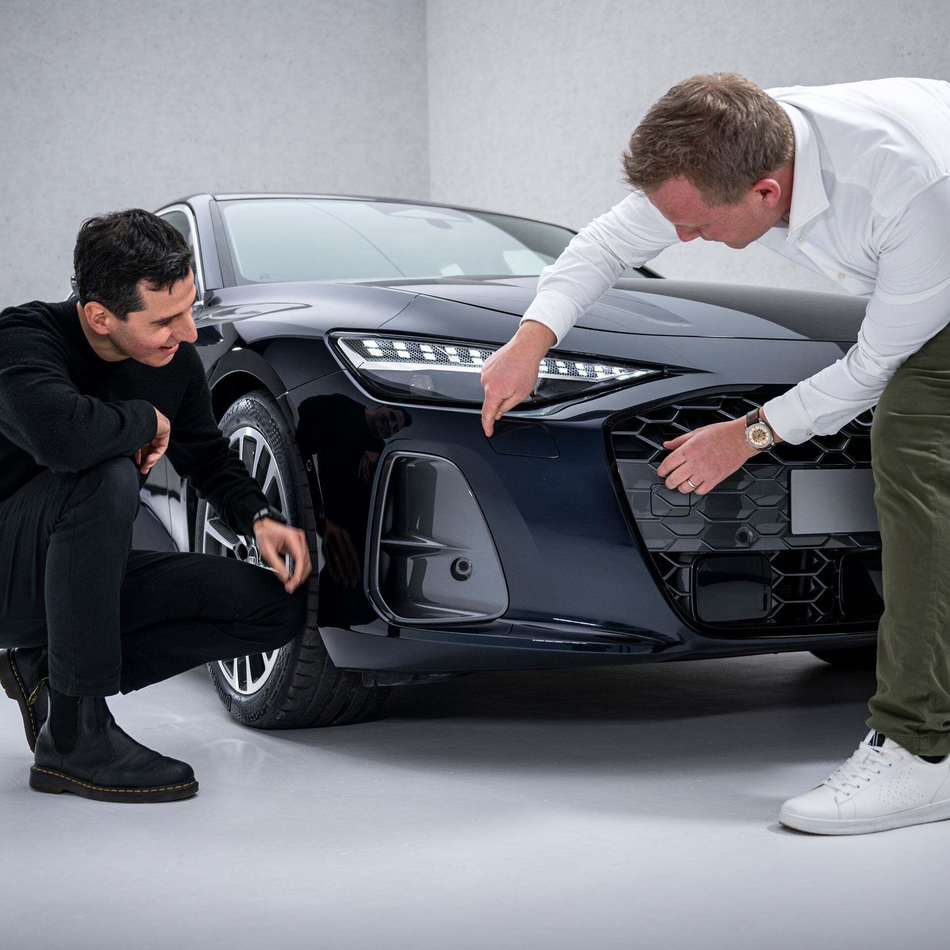 Francesco d’Amore (crouched down) and Sebastian Weiper (bent over) examine the front end of a black Audi A6 Sedan, specifically looking at the side air intakes, the so-called air curtains.