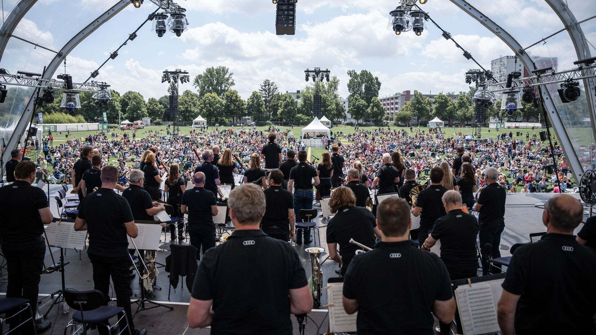 Orchester auf Open-Air-Bühne spielt vor großem Publikum im Park bei sonnigem Wetter.