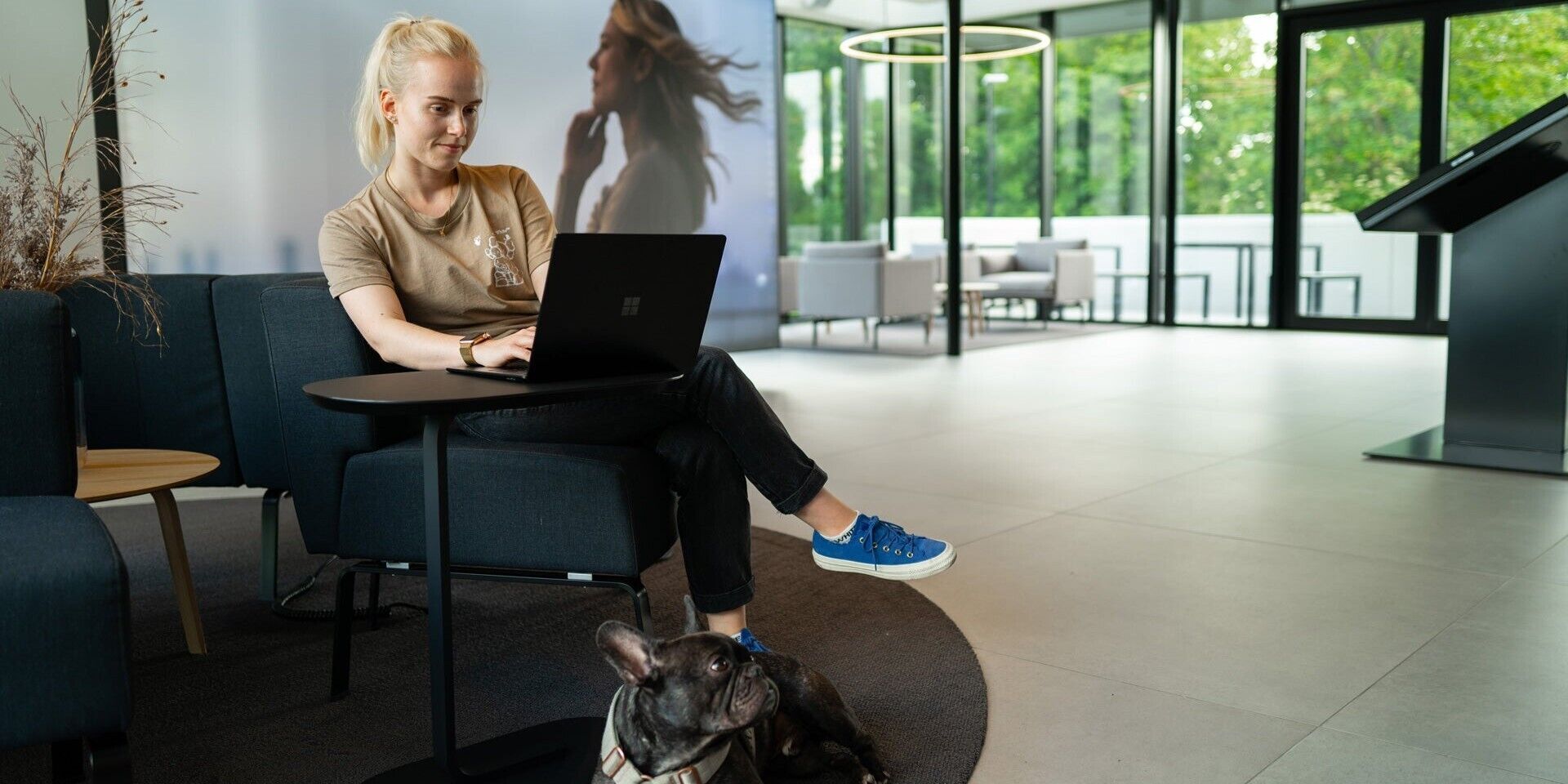 A woman sits in the charging hub at her laptop with her dog under the table