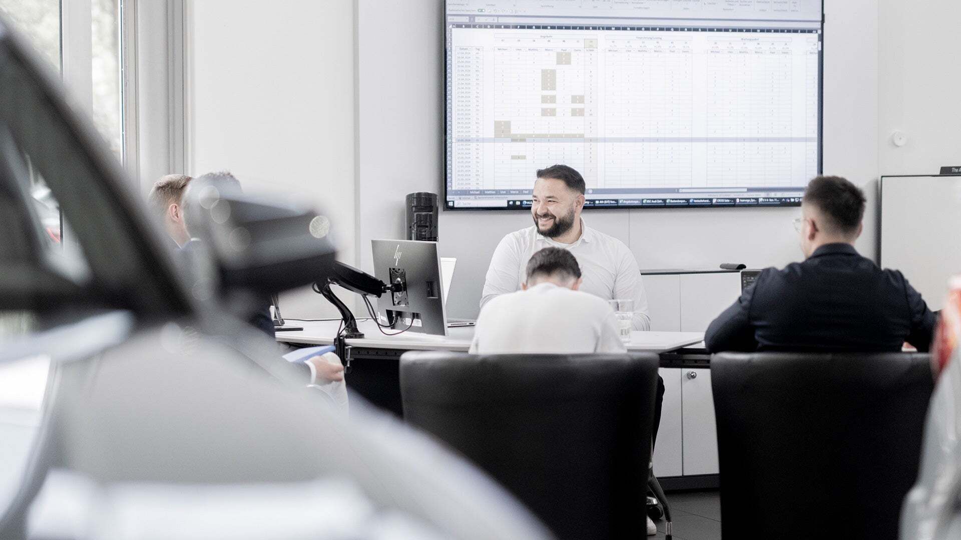 People are sitting at a table and talking. A screen with a document on it hangs on the wall.