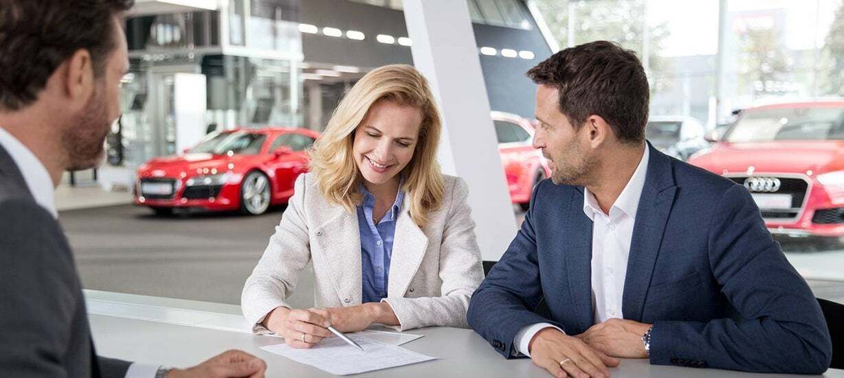 Drei Personen an einem Tisch in einem Autohaus, eine Frau unterschreibt ein Dokument; im Hintergrund rote Audi-Fahrzeuge.