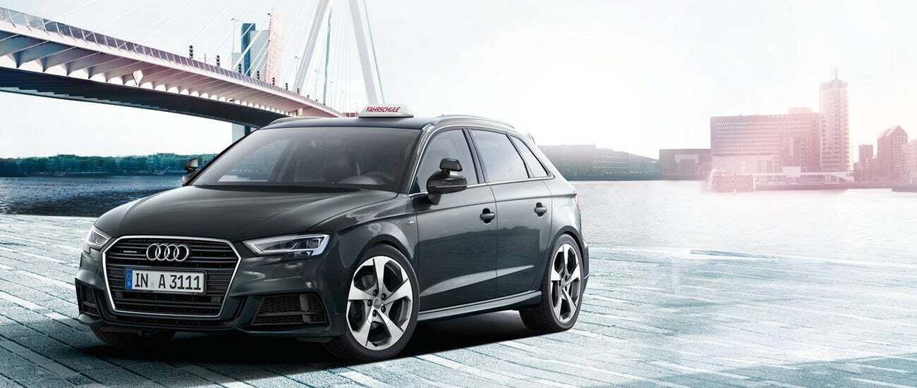Grauer Audi A3, geparkt an einer Uferpromenade mit einer Brücke im Hintergrund und einem Schild mit der Aufschrift „Fahrschule” darüber.