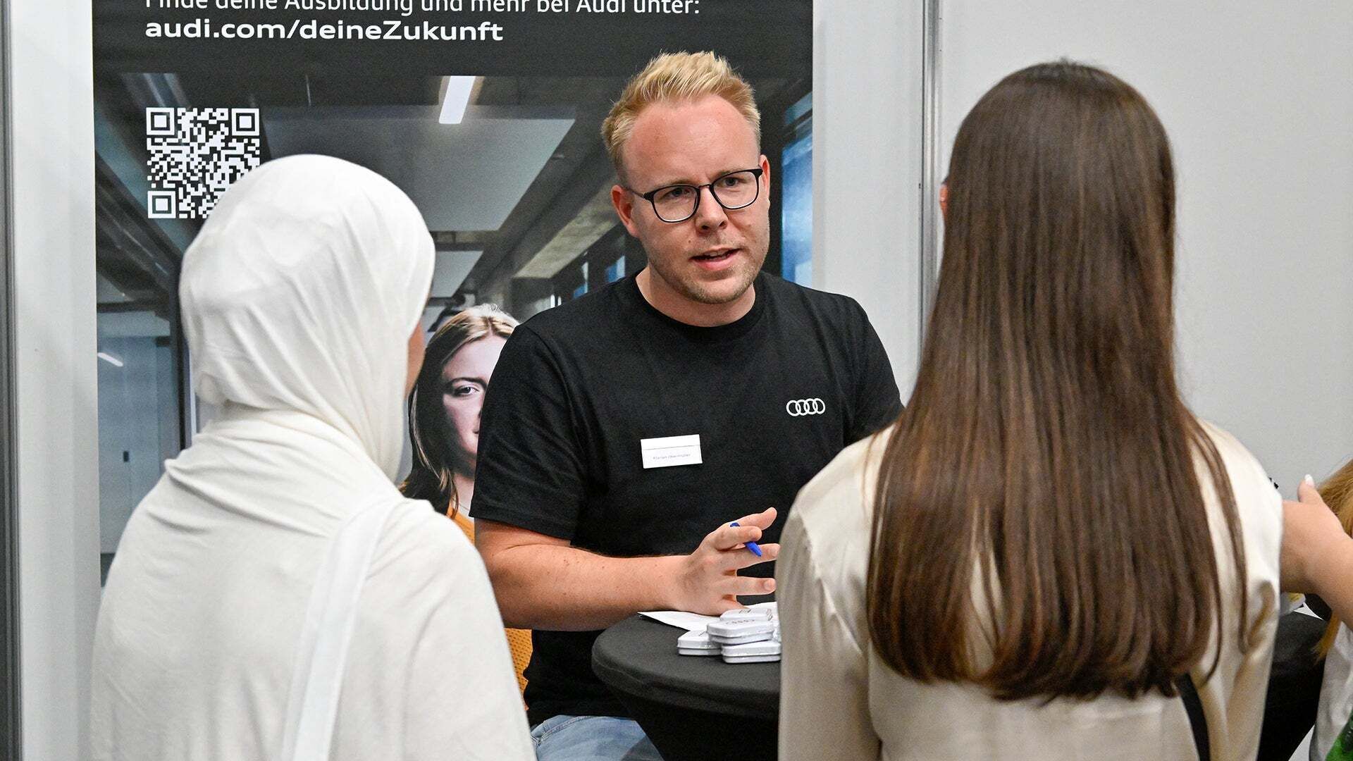 Ein Mann im schwarzen T-Shirt mit Audi Logo berät zwei Interessentinnen auf der Messe.