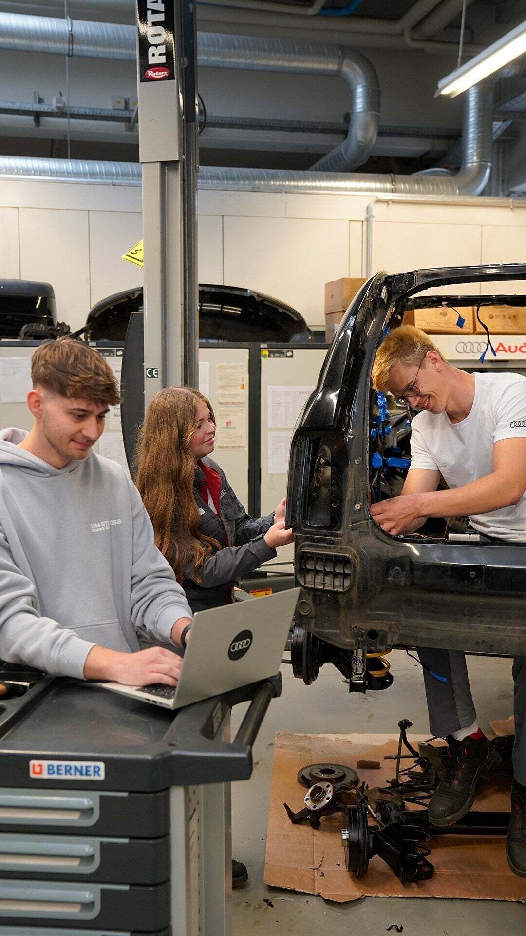 Zwei Auszubildende verrichten Arbeiten an einer Auto-Karosserie und ein anderer ist am Laptop
