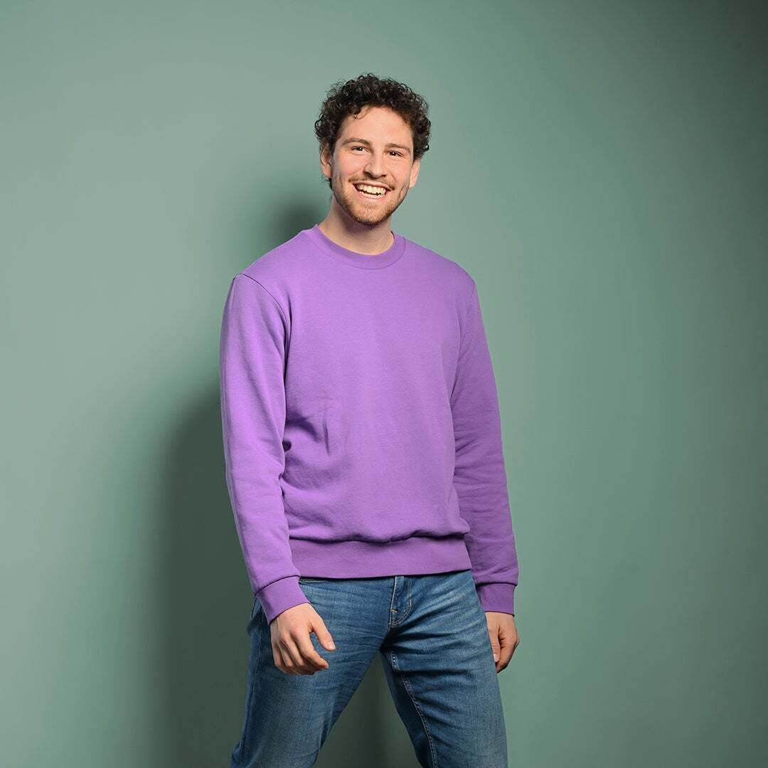 Fabian schaut in die Kamera und lacht. Er trägt einen lilanen Pullover