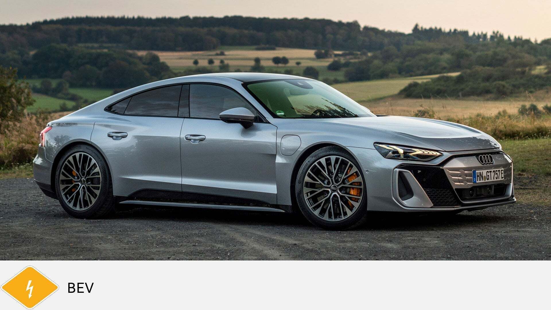 Silberner Audi RS e-tron GT steht auf einem Feldweg vor Wiese und Wald. Unten gelbes BEV-Symbol mit Blitz.