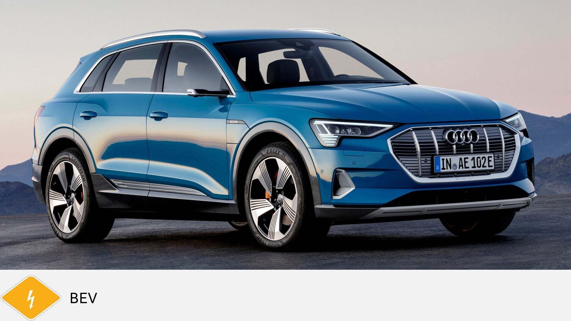 Blauer Audi e-tron SUV steht auf Asphalt vor Bergpanorama, schräg von vorne. Unten gelbes BEV-Symbol mit Blitz.