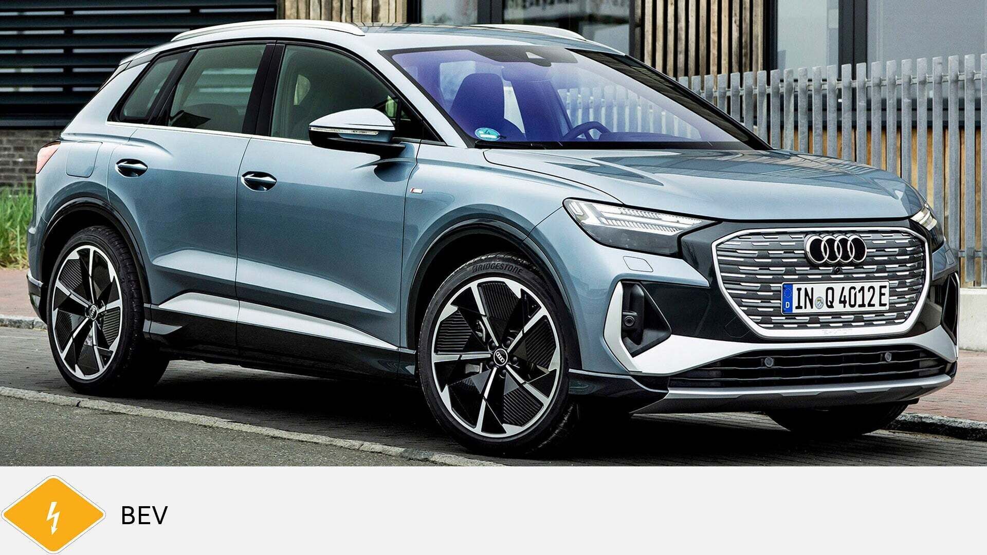 Grauer Audi Q4 e-tron SUV steht vor Gebäude mit Holz- und Metallfassade. Unten gelbes BEV-Symbol mit Blitz.