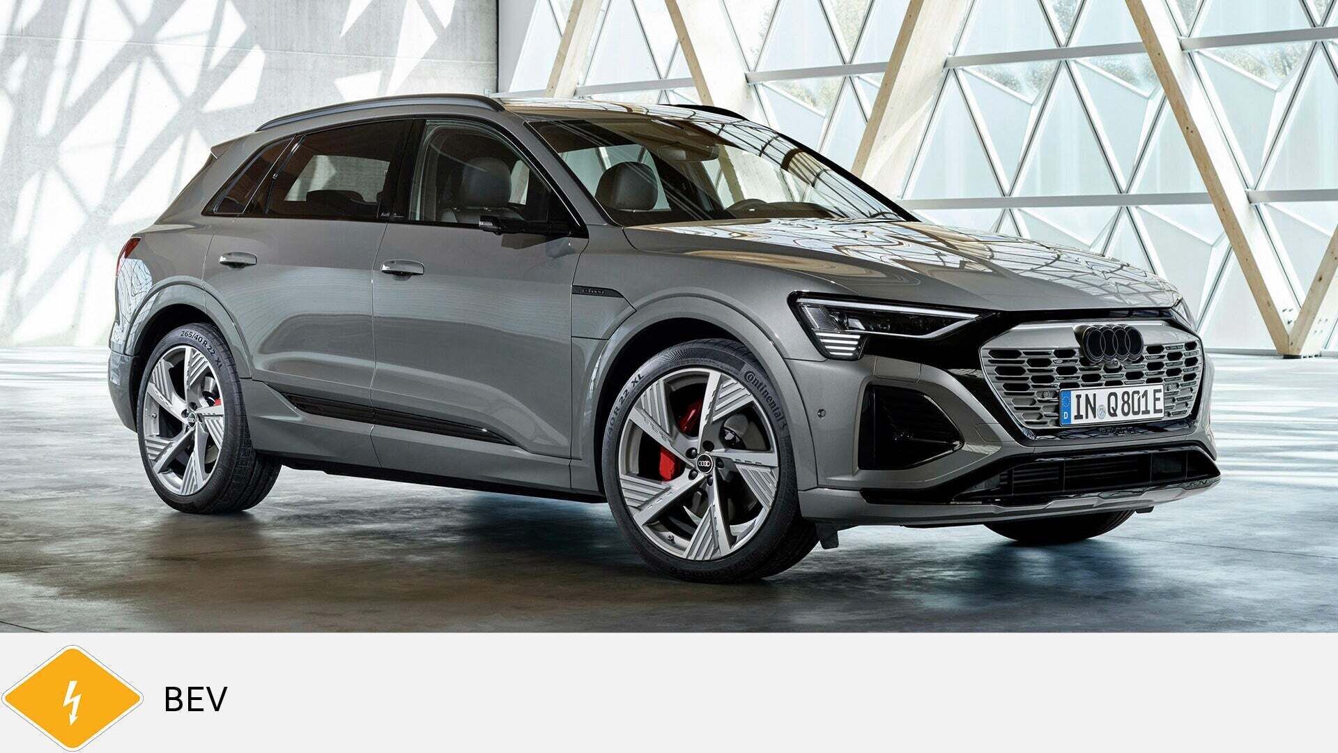 Grauer Audi Q8 e-tron SUV steht in lichtdurchflutetem Raum mit Glasstruktur. Unten gelbes BEV-Symbol mit Blitz.