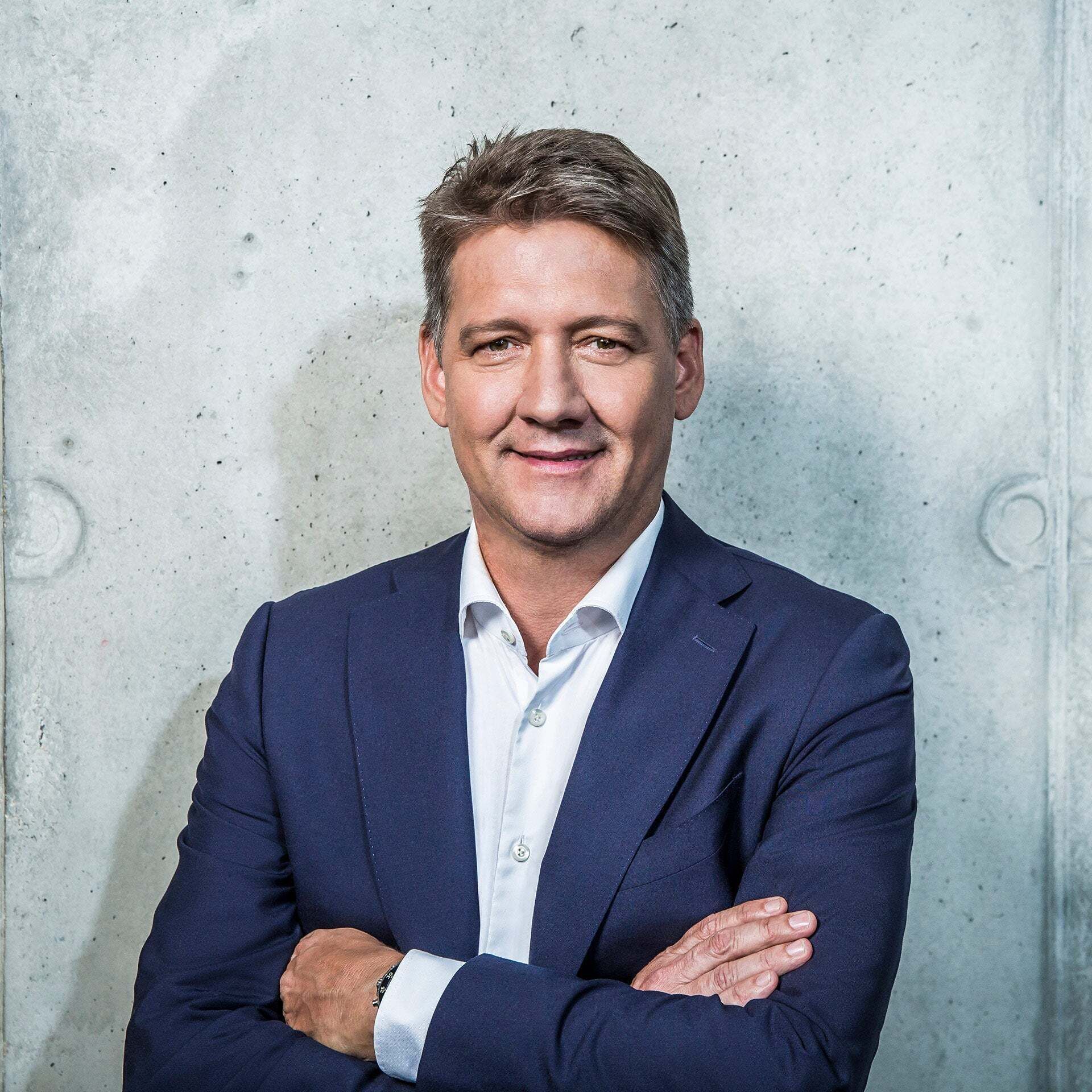 Gernot Döller stands with his arms crossed in front of a gray wall and smiles.