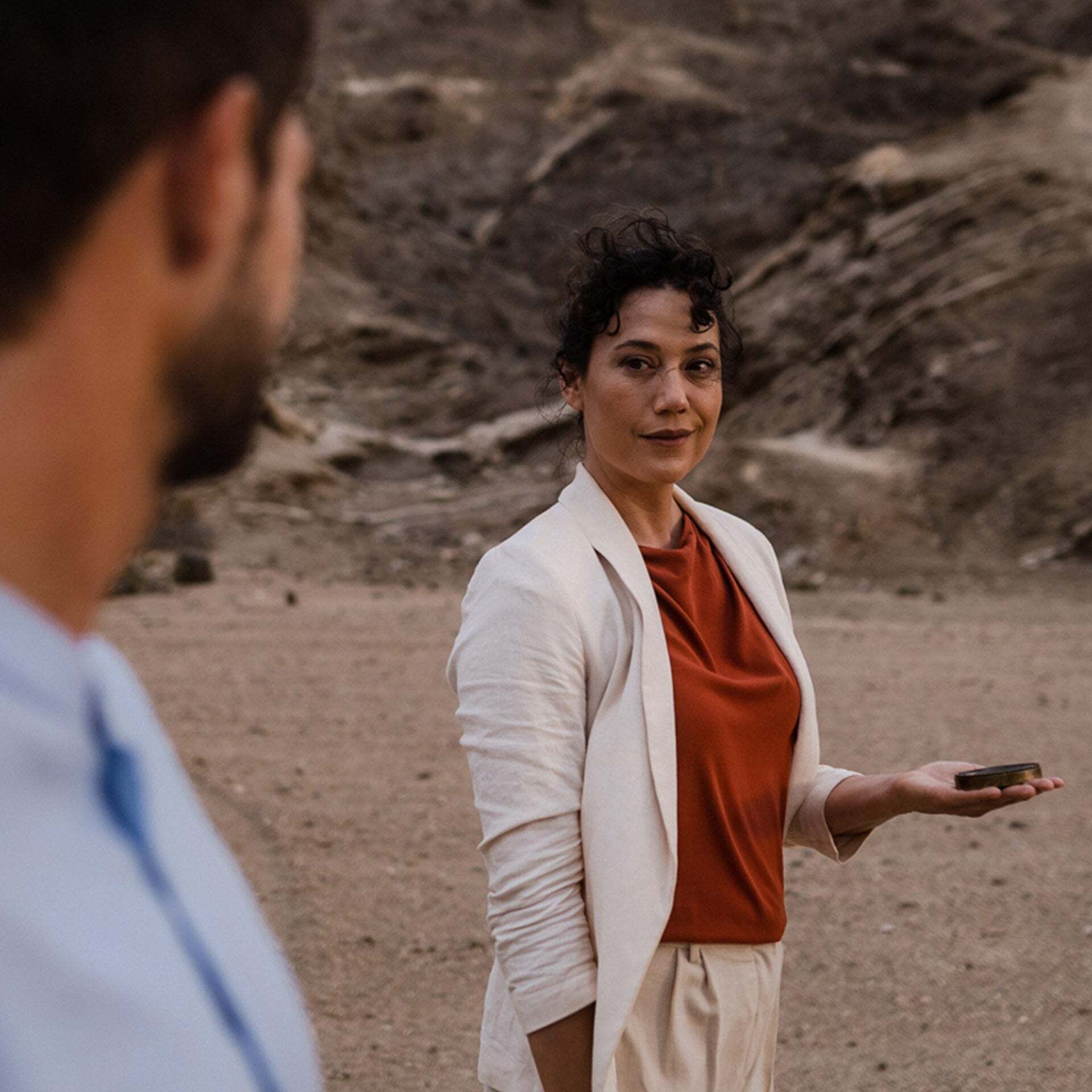 A woman stands in a desert landscape and looks into the eyes of a man in the foreground. A compass lies on her outstretched left hand.