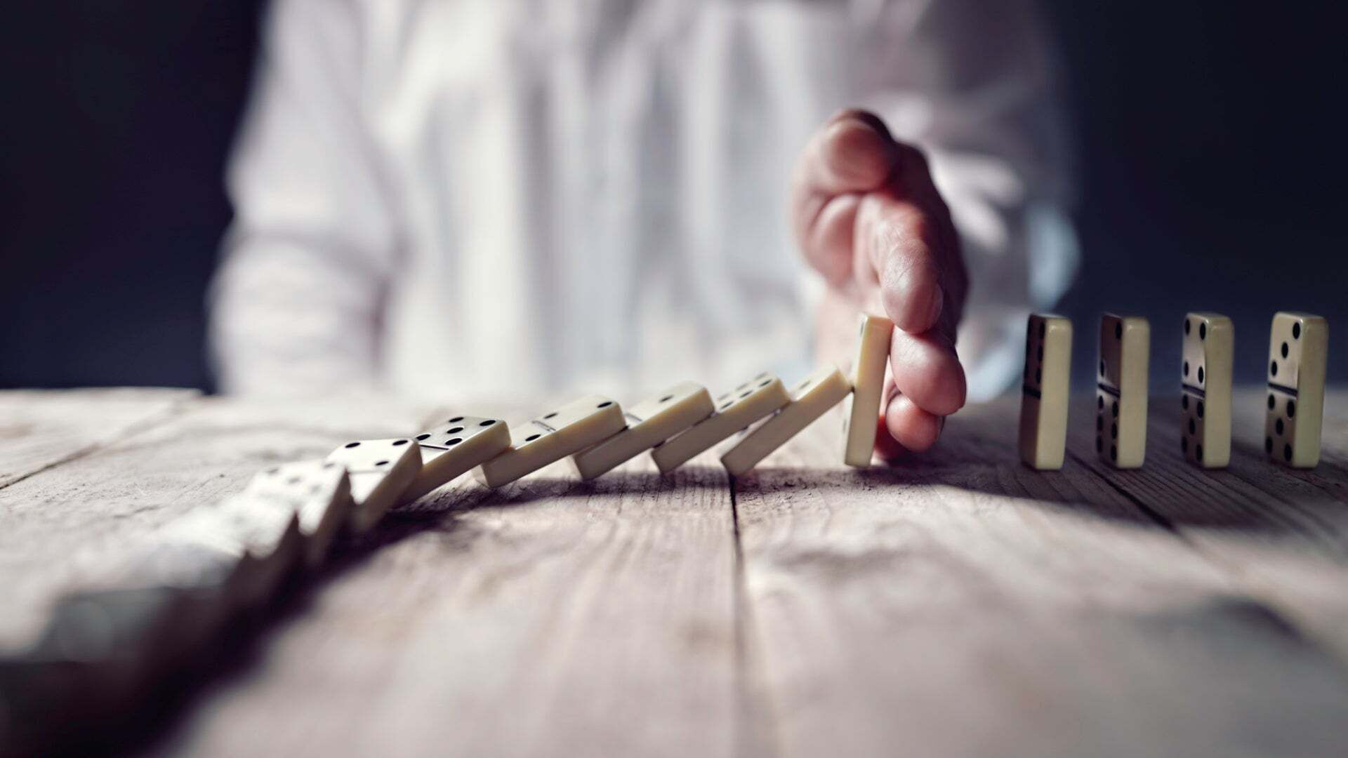 Ein Mann hält seine Hand auf einen Tisch, um umfallende Dominosteine zu stoppen.