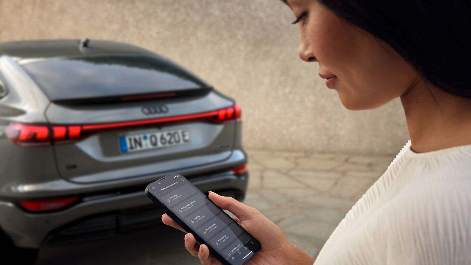 Eine Frau schaut auf ihr Handy mit der myAudi App. Im Hintergrund steht ein Audi Q6