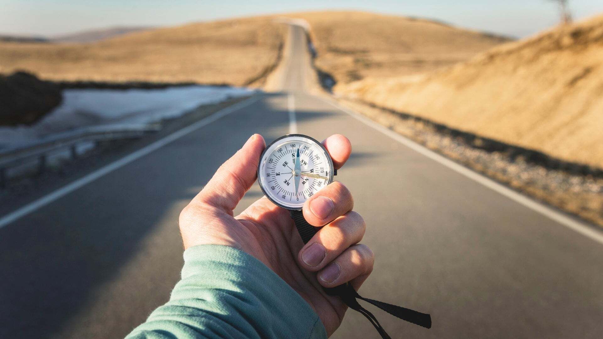 Eine Hand hält einen Kompass auf einer leeren, geraden Landstraße, die durch eine hügelige, wüstenähnliche Landschaft führt. - Eine Hand hält einen Kompass auf einer leeren, geraden Landstraße, die durch eine hügelige, wüstenähnliche Landschaft führt.
