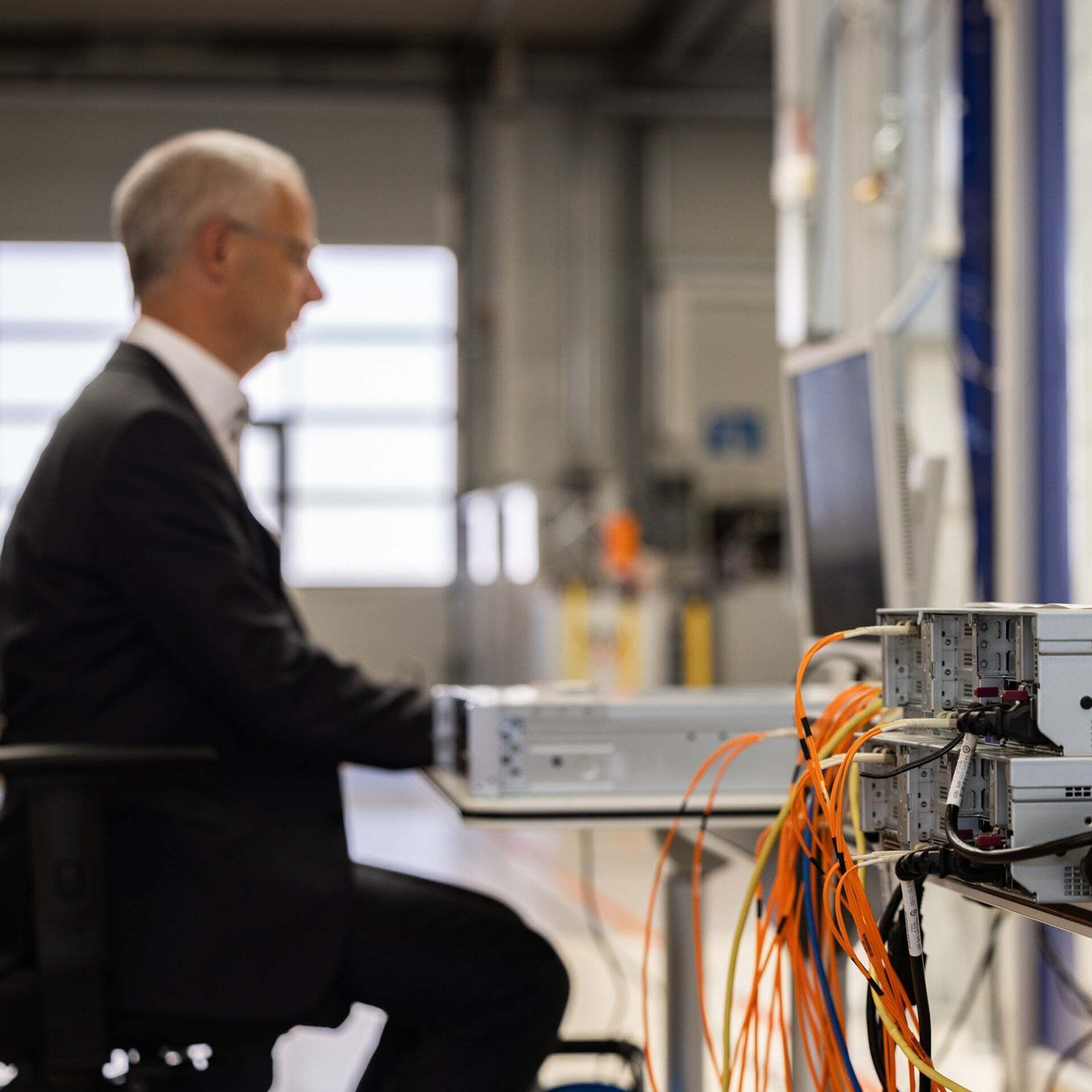 Component with orange cables protruding from it. In the background, Henning Löser is sitting at a computer workstation in the P-Lab.