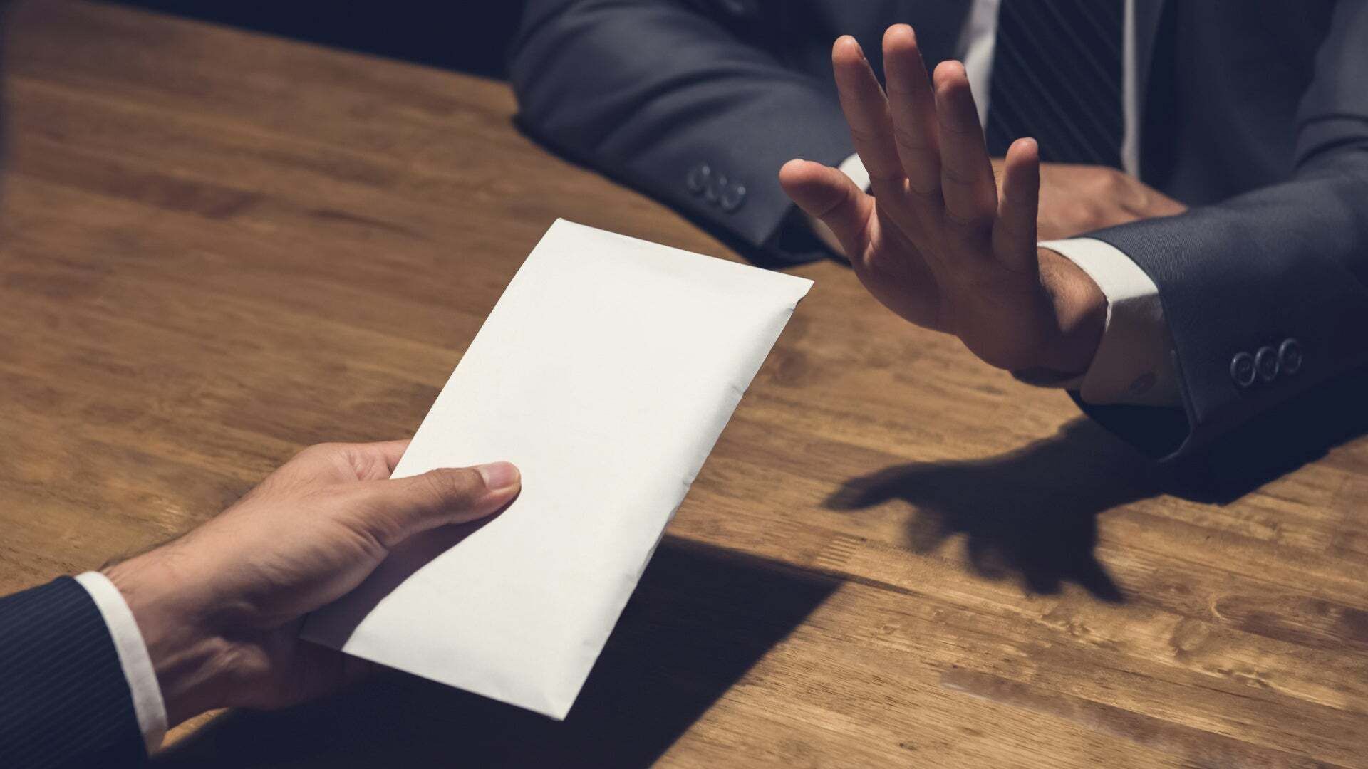 A hand passes an envelope across the table, the other person raises a hand in refusal.