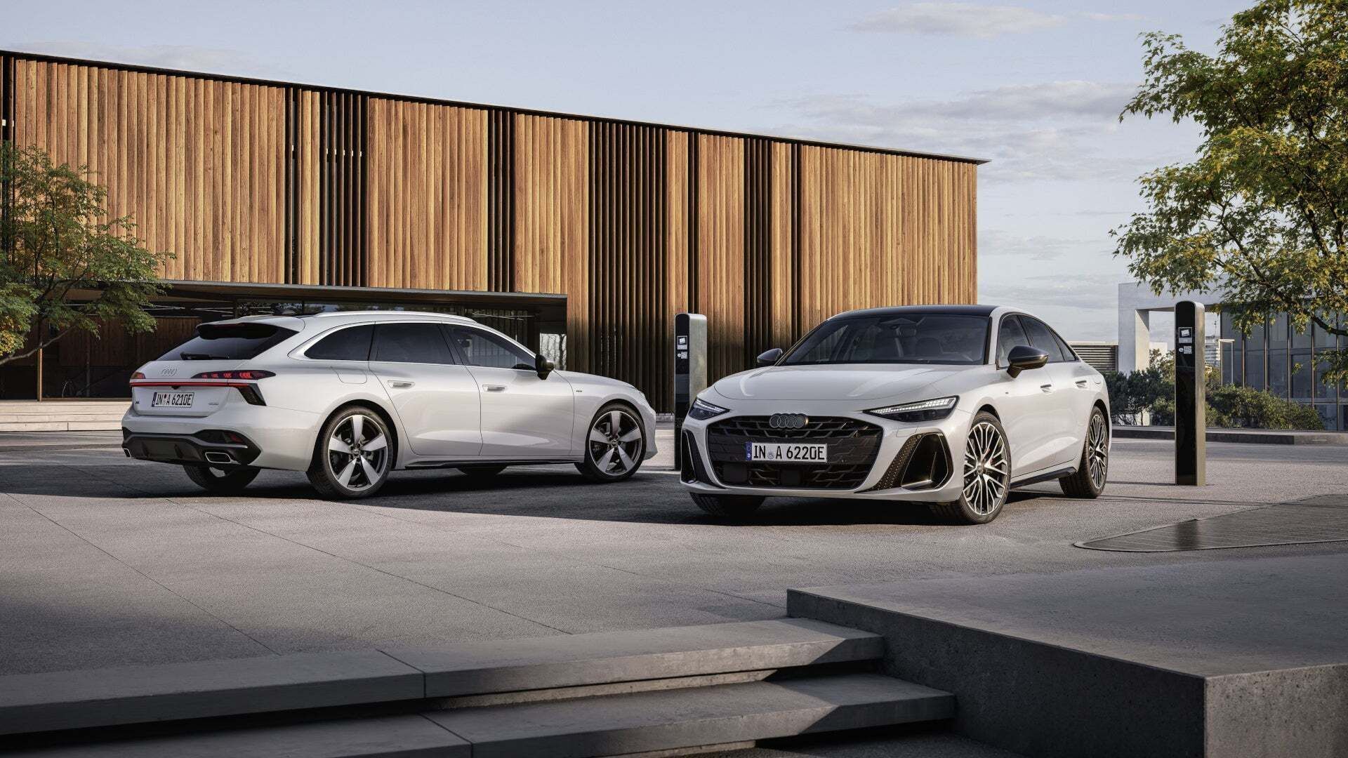 A white Audi A6 Avant e‑hybrid quattro and a white Audi A6 Sedan e‑hybrid quattro are parked in front of a modern building and connected to electric vehicle charging stations.