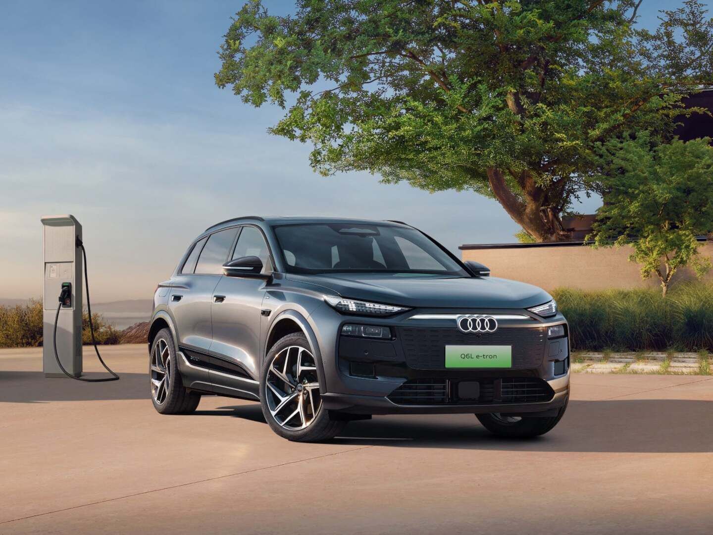 A grey‑blue Audi Q6L e‑tron is parked at a charging station in an open landscape under warm sunlight.