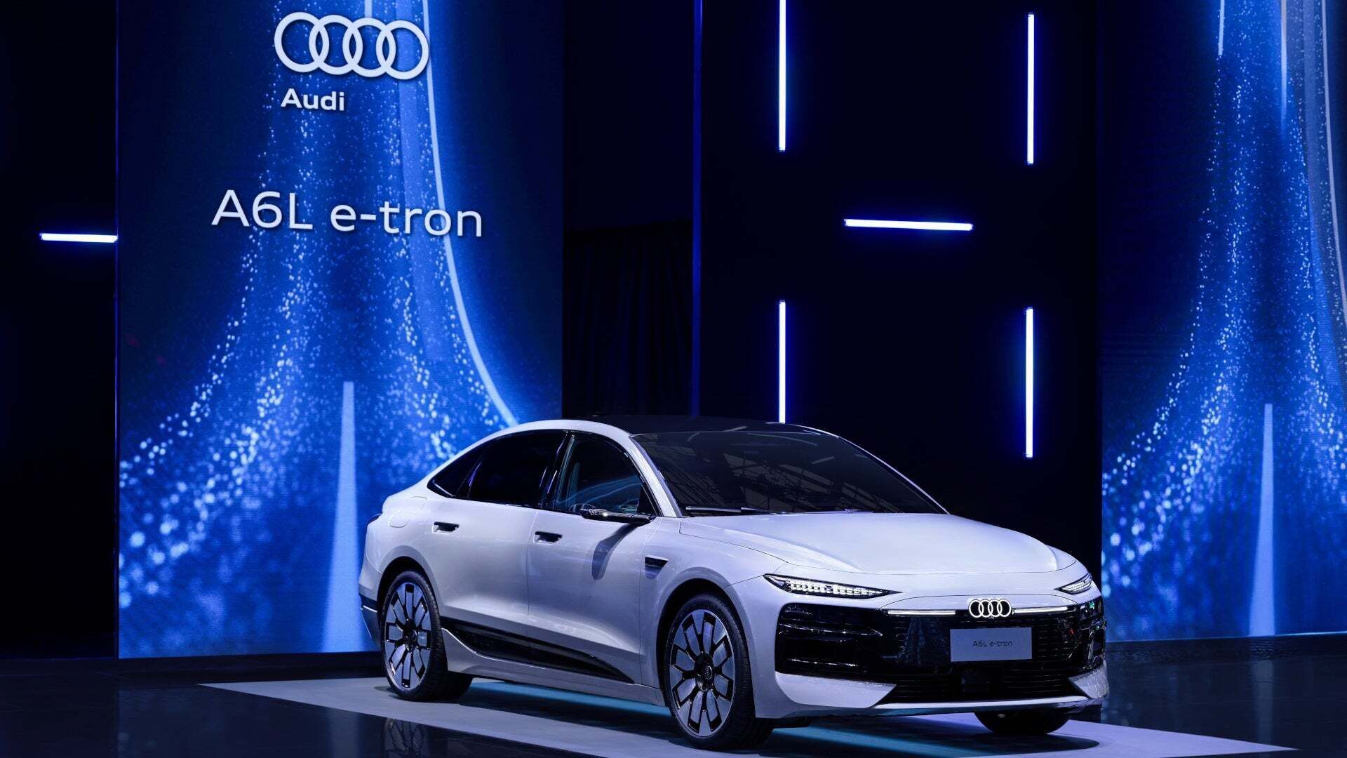 A white Audi A6L e‑tron is displayed on a stage in front of blue‑lit panels featuring the lettering ‘A6L e‑tron’ and the Audi rings.